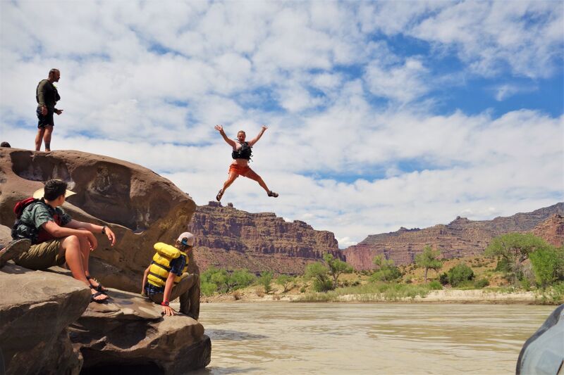 A group of people are enjoying a sunny day by a river. One person is captured mid-air, leaping from a rocky outcrop into the water with arms outstretched. Others are watching, some sitting on the rocks and another standing at the edge. The landscape features a wide river, rocky formations, and distant hills under a partly cloudy sky. The overall scene suggests a recreational activity like cliff jumping or swimming.